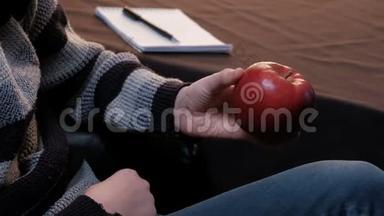 一个穿条纹毛衣的年轻人，手里拿着一个红苹果。 一个少年从桌子上拿起一个记事本和一支笔